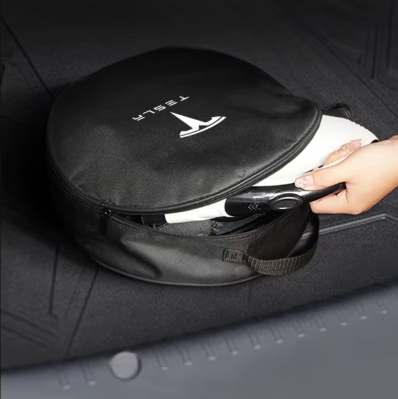 Sac noir circulaire Tesla avec logo et inscription 'TESLA' imprimés en blanc sur le dessus. Le sac est partiellement ouvert, laissant entrevoir un câble de recharge Tesla noir et blanc, tenu par une main. Le sac est posé sur une surface sombre, probablement le coffre d'une voiture