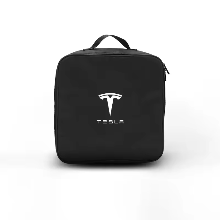 Sac noir carré Tesla avec logo et inscription 'TESLA' imprimés en blanc sur le dessus. Le sac est partiellement ouvert, laissant entrevoir un câble de recharge Tesla noir et blanc, tenu par une main. Le sac est posé sur une surface sombre, probablement le coffre d'une voiture